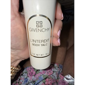 Puede incluir: Un envase blanco de talco corporal Givenchy L'Interdit. La etiqueta presenta el logotipo de Givenchy y los detalles del producto. El peso neto es de 56 g. El envase se sostiene en una mano.