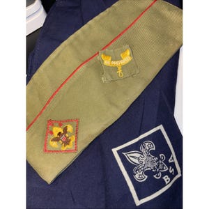 May include: An olive green Boy Scout hat with a red stripe and two patches. One patch reads "BE PREPARED" and the other features an eagle. A navy blue neckerchief with a BSA emblem is also visible.