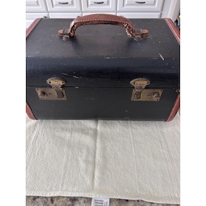 May include: A vintage black suitcase with brown leather trim and handle. The suitcase has two brass latches and is sitting on a white cloth. The suitcase appears to be well-worn.