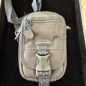 Può includere: Una borsa a tracolla grigia Samsonite con cinturino nero e dettagli blu. La borsa ha una piccola tasca frontale, una chiusura a fibbia e il logo Samsonite. Il cinturino è regolabile e presenta dettagli blu a pois.