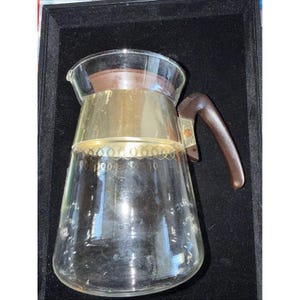 May include: A clear glass coffee carafe with a brown plastic handle and lid. The carafe has a gold-colored metal band around the middle with a decorative pattern. The carafe is empty and sits on a black surface.