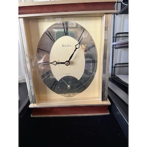 May include: A rectangular Bulova quartz clock with a gold-colored face and silver-colored frame. The clock face has black Roman numerals and hands. The clock has a small, round dial at the bottom. The clock is set on a brown base.