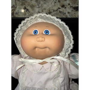 May include: A vintage Cabbage Patch Kids doll with a light skin tone, blue eyes, and a small, pursed mouth. The doll wears a pink dress with a white lace collar and a matching bonnet. A white ribbon is tied around the neck.
