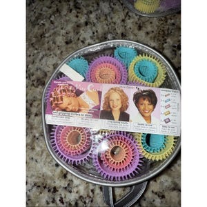 May include: A clear, round case containing self-gripping hair curlers in various colors. The curlers are in shades of pink, purple, teal, and yellow. The packaging includes text that reads "self-gripping curlers to sleep" and "Olivia Garden".