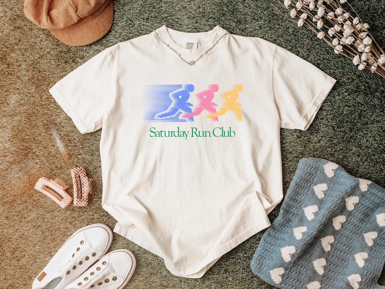 May include: Cream-colored t-shirt with a graphic of three runners in blue, pink, and yellow, with the text "Saturday Run Club." The shirt is paired with white sneakers, a brown cap, and a blue blanket with white hearts.