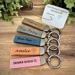 Könnte beinhalten: Fünf rechteckige Schlüsselanhänger aus Leder in Beige, Grau, Blau, Orange und Pink, jeweils mit einem dunklen Metallverschluss und Ring. Die Anhänger sind graviert mit den Texten Mama mit Herz, Für Papa, Bester Papa mit Herz, Amelie mit Herz und DRAMA QUEEN mit Kronensymbol.