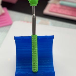 May include: A green hand gesture on a silver telescoping wand, resting in a blue holder. The wand's handle has a textured green grip. The hand is in a 'peace' or 'victory' sign.