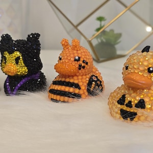 May include: Four rubber ducks decorated with small rhinestones. The first is brown and white like a football, the second is black and yellow, the third is orange and black with bee stripes, and the fourth is yellow with a black jack-o'-lantern face.