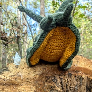May include: A crochet snail plush toy featuring a dark green shell and a textured mustard yellow underside. The snail has two long upper tentacles and two shorter lower tentacles. A small white gnome figurine stands to the left of the snail.