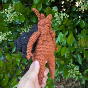 May include: A handmade crochet snail toy featuring a textured, rust-colored body with curled antennae and a large, dark charcoal-colored spiral shell. The yarn work displays a dense, uniform stitch pattern throughout the entire figure.