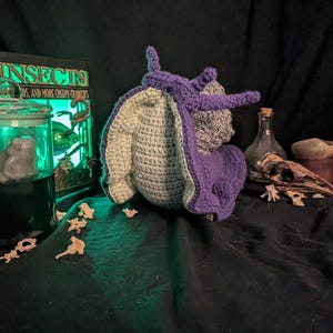 May include: A crocheted snail plush featuring a vibrant purple body and shell with a light gray underside. The snail is positioned next to a glass jar containing a gray figure, a book titled INSECTS, a glass bottle, and several small bone-like pieces.