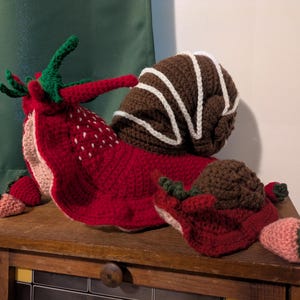 May include: A large crocheted snail with a red strawberry-patterned body, green leafy antennae, and a brown spiral shell featuring white stitched accents. A smaller matching crocheted snail sits beside it, both decorated with small pink and red strawberry shapes.