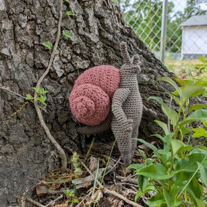 May include: A crocheted snail figurine featuring a textured, dusty rose-colored spiral shell attached to a brown, anthropomorphic body with two upright antennae and small arms, crafted with visible yarn stitches throughout the entire surface.