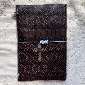 May include: A dark brown journal with a textured snakeskin pattern, secured by a light blue elastic band. The band features two small white and blue floral beads and a silver-toned metal cross charm hanging from the center of the cover.