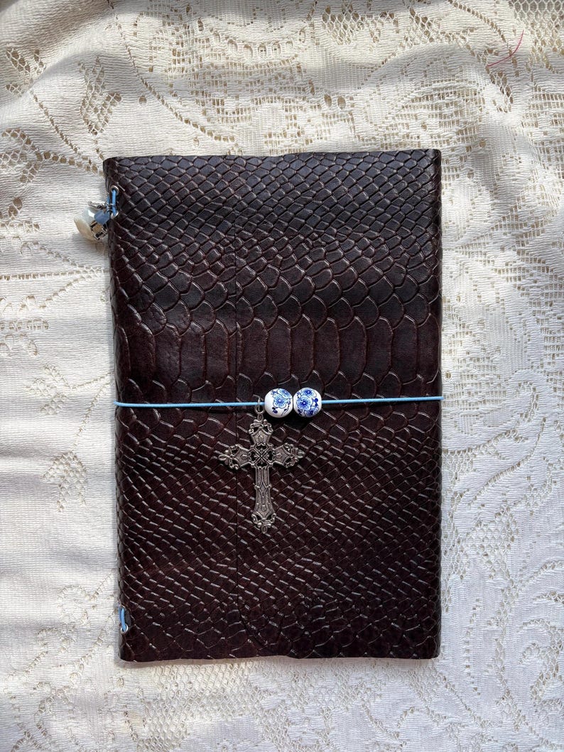 Puede incluir: Un diario de color marr&oacute;n oscuro con un patr&oacute;n texturizado de piel de serpiente, asegurado por una banda el&aacute;stica azul claro. La banda presenta dos peque&ntilde;as cuentas blancas y azules con motivos florales y un dije de cruz plateada.