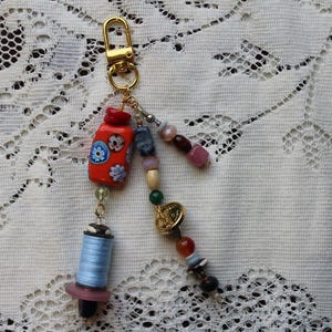 Op de afbeelding: Een handgemaakte sleutelhanger met een gouden sluiting, met een decoratieve flesbedel in rood, oranje en blauw, een blauwe garenspoel en diverse kleurrijke kralen. Een uniek accessoire.