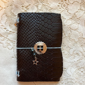 May include: A dark brown notebook with a textured snakeskin pattern. The cover is secured by a light blue elastic band featuring a silver-toned circular concho ornament, a small dangling star charm, and dark beads attached to the elastic cord.