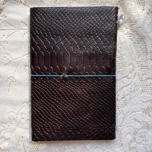 Puede incluir: Un cuaderno rectangular con una cubierta de color marr&oacute;n oscuro que presenta un patr&oacute;n texturizado y en relieve de piel de serpiente. El cuaderno est&aacute; asegurado por una banda el&aacute;stica delgada de color azul claro envuelta horizontalmente.