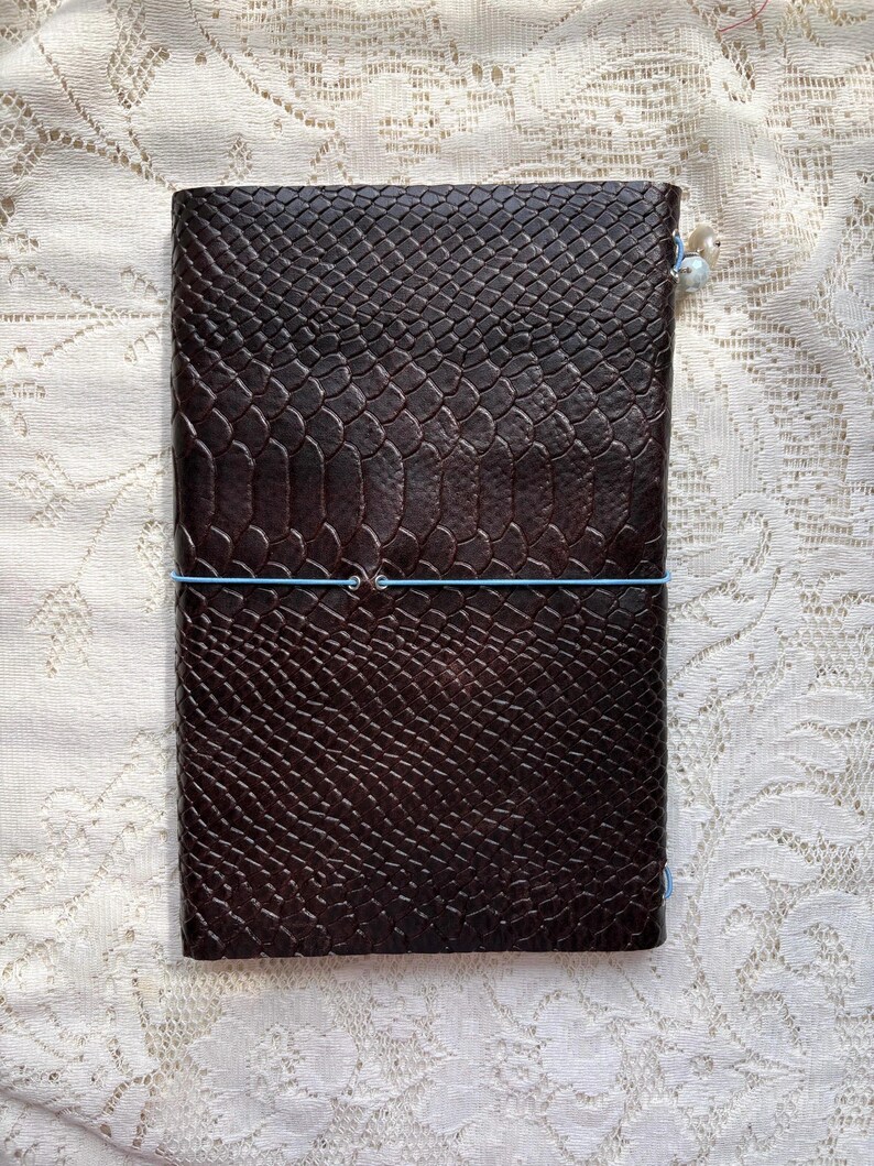 Puede incluir: Un cuaderno rectangular con una cubierta de color marr&oacute;n oscuro que presenta un patr&oacute;n texturizado y en relieve de piel de serpiente. El cuaderno est&aacute; asegurado por una banda el&aacute;stica delgada de color azul claro envuelta horizontalmente.