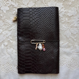 May include: A dark brown notebook cover with a textured snakeskin pattern. A gold-toned safety pin is attached to the front, featuring a cluster of small dangling beads in shades of white, pink, and orange, secured by a thin black elastic band.