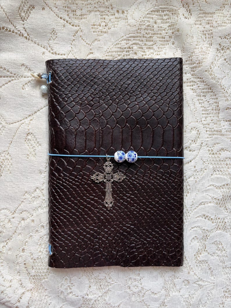 Puede incluir: Un diario de color marr&oacute;n oscuro con un patr&oacute;n texturizado de piel de serpiente. Una banda el&aacute;stica azul claro envuelve horizontalmente la cubierta, con dos peque&ntilde;as cuentas florales blancas y azules y un dije de cruz de metal plateado.