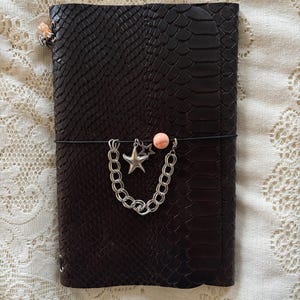 May include: A dark brown notebook with a textured snakeskin pattern cover. A black elastic band wraps around the center, holding a silver-toned metal chain, a star-shaped charm, and a small, round, peach-colored bead as decorative accents.