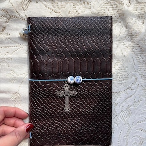 Puede incluir: Un diario rectangular de color marr&oacute;n oscuro con un patr&oacute;n texturizado similar a la piel de serpiente. Una banda el&aacute;stica azul claro envuelve horizontalmente la cubierta, con dos peque&ntilde;as cuentas redondas blancas y azules y una cruz plateada.