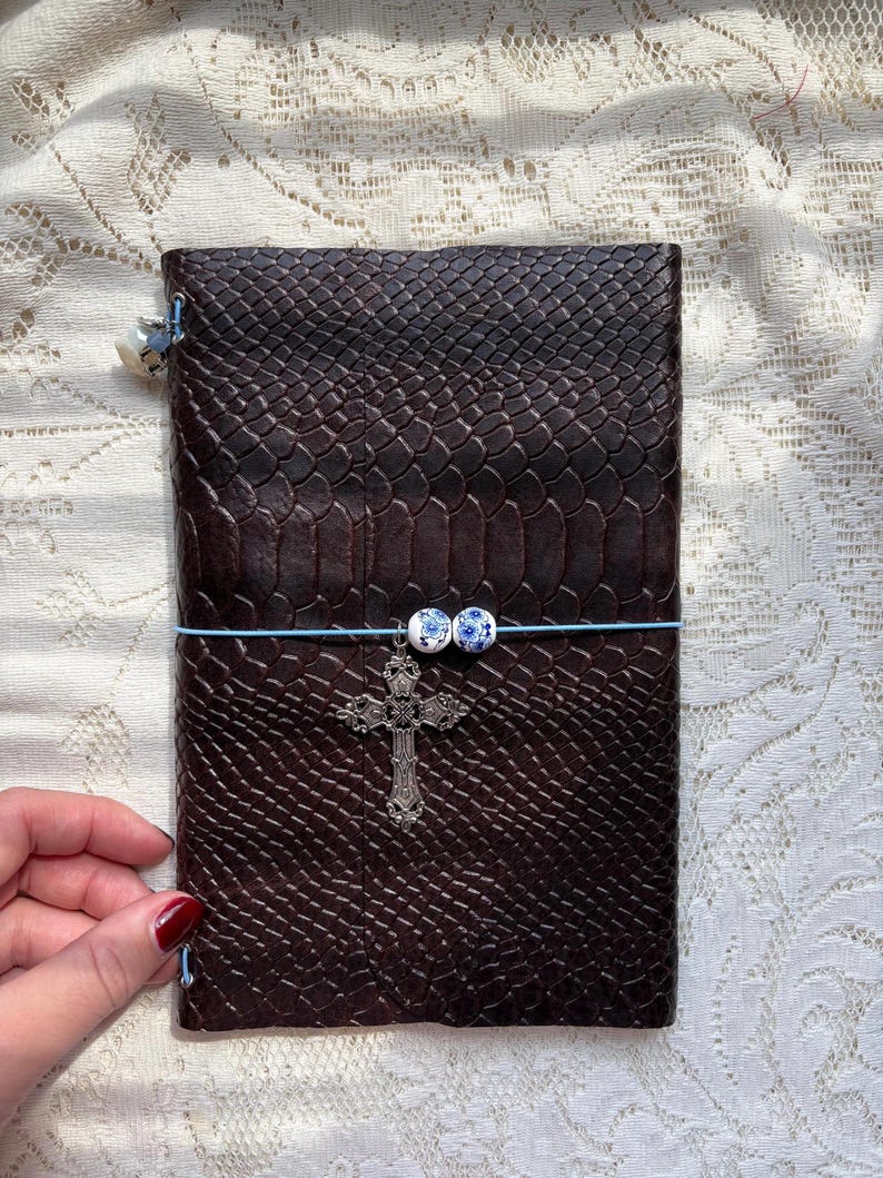 Puede incluir: Un diario rectangular de color marr&oacute;n oscuro con un patr&oacute;n texturizado similar a la piel de serpiente. Una banda el&aacute;stica azul claro envuelve horizontalmente la cubierta, con dos peque&ntilde;as cuentas redondas blancas y azules y una cruz plateada.