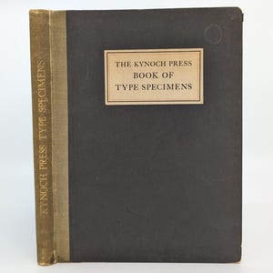 Può includere: Un libro con copertina rigida intitolato The Kynoch Press Book of Type Specimens con una copertina grigio antracite scuro e un dorso in tessuto color marrone chiaro. Il titolo è stampato in nero su un'etichetta rettangolare sul davanti e sul dorso.