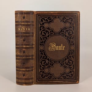 May include: A vintage book with a brown leather cover. The spine reads "DANTE" in gold lettering. The front cover features an ornate design with the name "Dante" in gold. The book's edges are worn, suggesting age.
