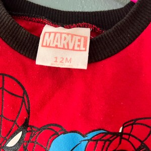 K&ouml;nnte beinhalten: Ein rotes T-Shirt mit schwarzem Rippkragen und einem Spider-Man-Motiv. Das wei&szlig;e Innenetikett zeigt den Text MARVEL und 12M. Das Kleidungsst&uuml;ck h&auml;ngt an einem leuchtend pinken Kunststoffkleiderb&uuml;gel vor einer hellblauen Wand.