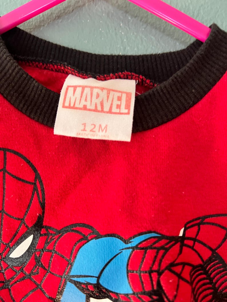 K&ouml;nnte beinhalten: Ein rotes T-Shirt mit schwarzem Rippkragen und einem Spider-Man-Motiv. Das wei&szlig;e Innenetikett zeigt den Text MARVEL und 12M. Das Kleidungsst&uuml;ck h&auml;ngt an einem leuchtend pinken Kunststoffkleiderb&uuml;gel vor einer hellblauen Wand.