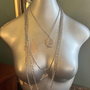 May include: Silver-toned layered necklaces displayed on a mannequin. The necklaces feature different chain styles, including a long chain with large links, a delicate chain, and a pendant necklace with a hammered coin-shaped charm.