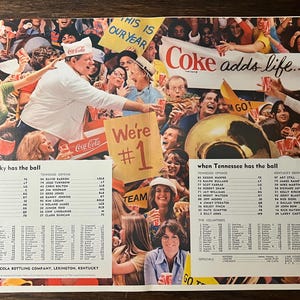 May include: Vintage Coca-Cola advertisement featuring a crowd of people at a sporting event. The ad includes the text "Coke adds life..." and "We're #1". People are holding Coca-Cola cups and wearing Coca-Cola hats.