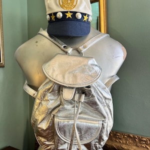 May include: A silver metallic backpack with a drawstring closure and a front pocket. The backpack is displayed on a mannequin with a white and navy blue hat with gold embellishments and star accents. The hat has a gold emblem.