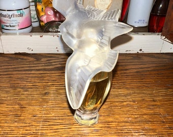 May include: A clear glass bottle of perfume with a white bird-shaped stopper. The bottle is sitting on a wooden shelf with other perfume bottles.