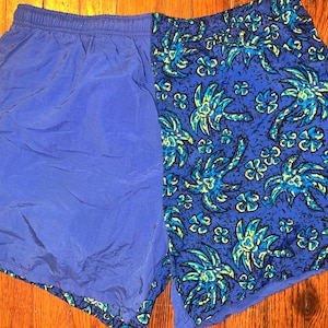 May include: Blue athletic shorts with a split design. One side is solid blue, the other features a blue and green tropical floral pattern. The shorts have an elastic waistband and are made of a lightweight material.