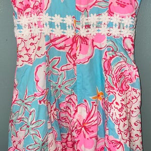 May include: A sleeveless dress featuring a vibrant floral pattern in shades of bright pink, light blue, and white. The garment includes a horizontal band of white crocheted daisy-like trim across the waist and a central vertical zipper closure.