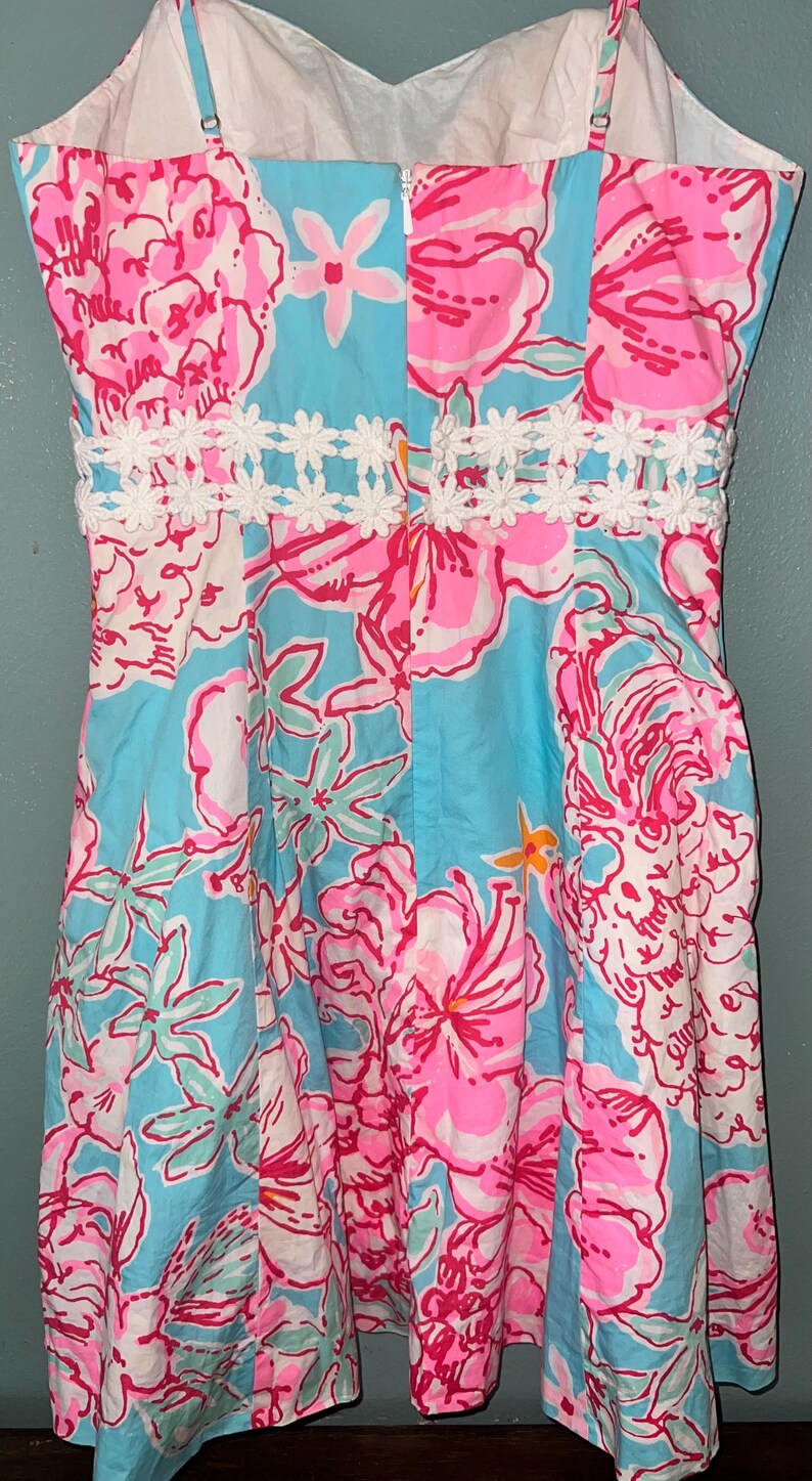 May include: A sleeveless dress featuring a vibrant floral pattern in shades of bright pink, light blue, and white. The garment includes a horizontal band of white crocheted daisy-like trim across the waist and a central vertical zipper closure.