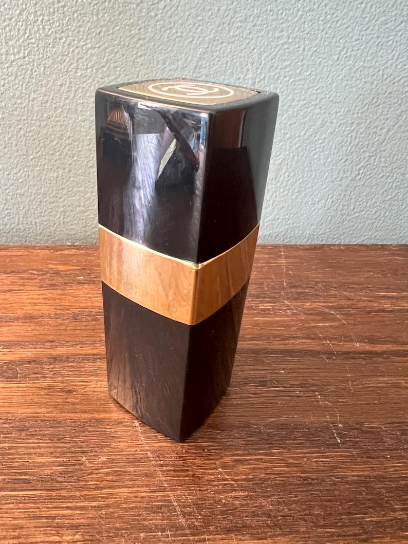 May include: A tall, rectangular black cosmetic container with a glossy finish. The object features a wide, horizontal gold-colored band encircling the middle section. The top cap displays a circular white logo with two interlocking C letters.