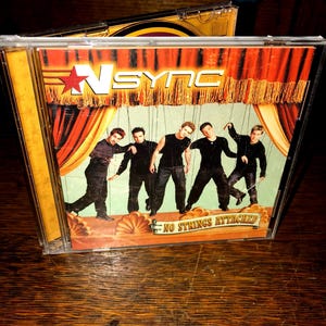 May include: A CD case with a red, white, and yellow design. The cover features a band called *NSYNC with the title "No Strings Attached".