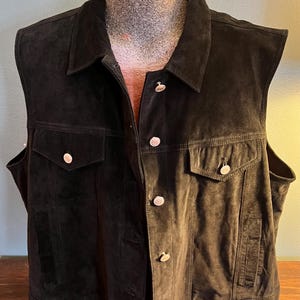 May include: A black suede vest with a collared neckline and two chest pockets. The vest has silver-colored buttons down the front and on the pockets. The vest is sleeveless and appears to be a medium size.