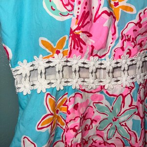 May include: A sleeveless dress featuring a vibrant floral print in shades of bright blue, hot pink, and yellow. The garment includes a horizontal cutout at the waist accented with a white, daisy-shaped lace trim that connects the bodice to the skirt.