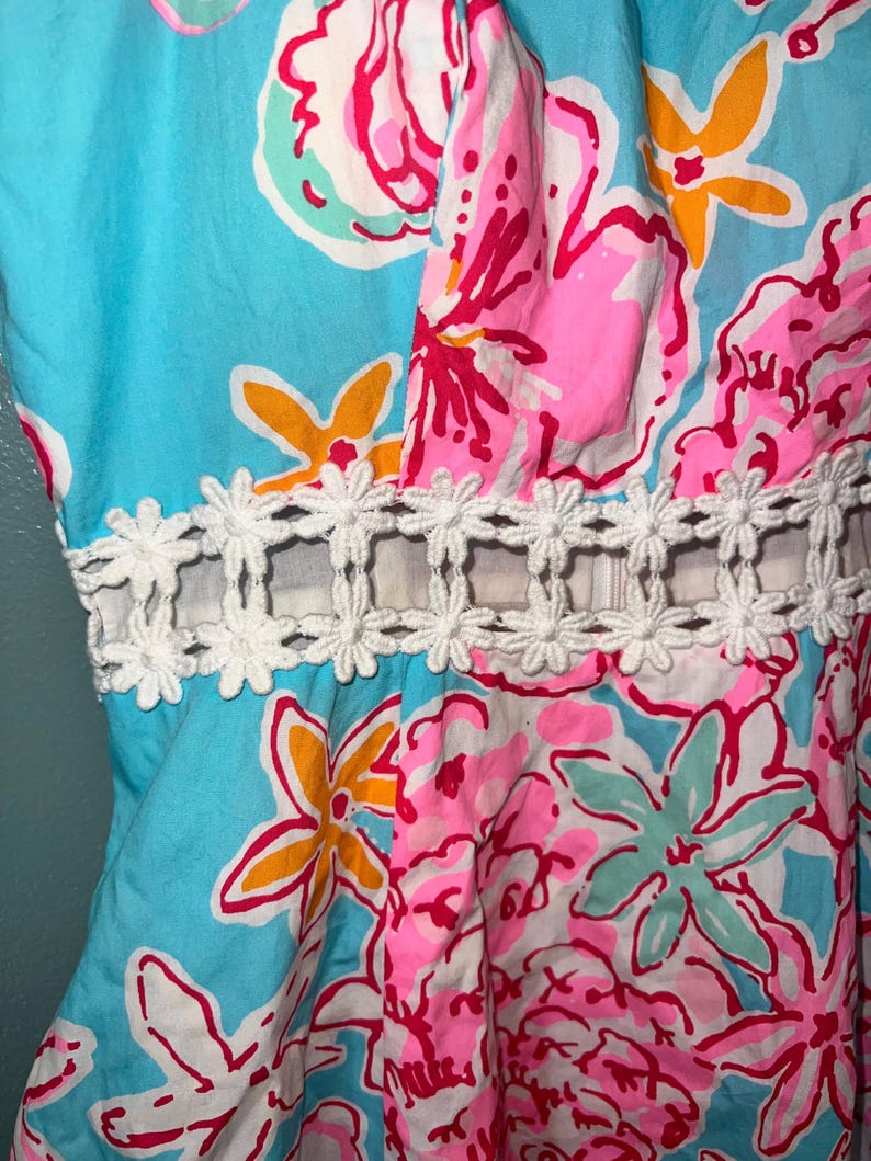 May include: A sleeveless dress featuring a vibrant floral print in shades of bright blue, hot pink, and yellow. The garment includes a horizontal cutout at the waist accented with a white, daisy-shaped lace trim that connects the bodice to the skirt.