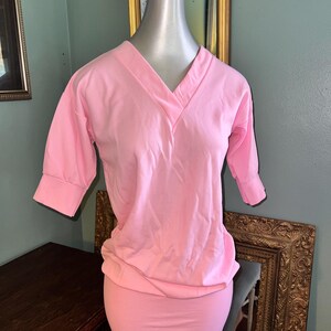May include: A light pink, V-neck top with three-quarter sleeves and a gathered waist. The top is displayed on a mannequin. The fabric appears to be a soft, lightweight material. The overall style is casual and feminine.