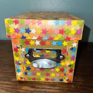 May include: A decorative box with a lid, covered in a pattern of colorful stars on a yellow background. The box has a metal label holder on the front and is secured with small metal rivets.