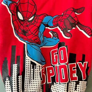 K&ouml;nnte beinhalten: Ein rotes Langarm-Sweatshirt mit einer Grafik der Figur Spider-Man in einer dynamischen Pose &uuml;ber einer schwarz-wei&szlig;en Stadtsilhouette. Der Text GO SPIDEY ist in fetten roten und wei&szlig;en Buchstaben aufgedruckt. Es hat einen schwarzen Rippkragen.