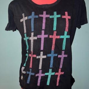 May include: A black short-sleeved t-shirt featuring a grid pattern of twenty crosses in various colors including light blue, purple, teal, pink, and white. The crosses have a distressed, weathered texture and are arranged in four rows of five.