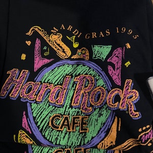 May include: Black Hard Rock Cafe t-shirt with a colourful graphic. The design includes the words "Mardi Gras 1995" and "Hard Rock Cafe" in a vibrant, retro style. A saxophone and musical notes are also featured.