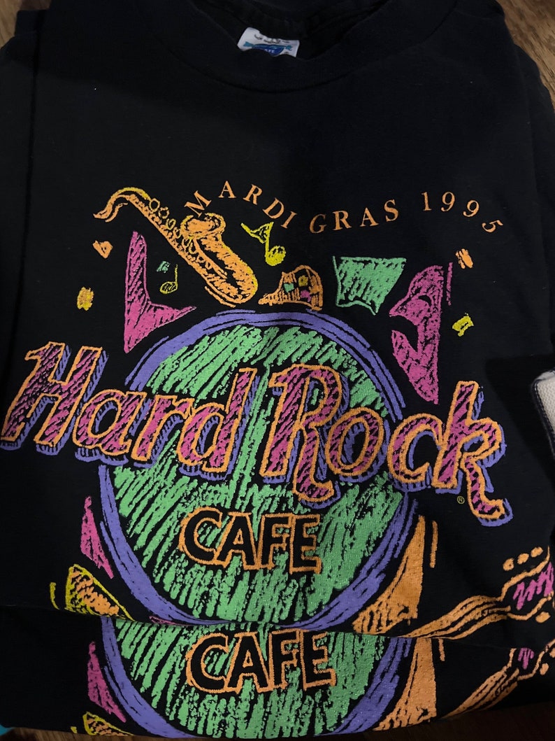 May include: Black Hard Rock Cafe t-shirt with a colourful graphic. The design includes the words "Mardi Gras 1995" and "Hard Rock Cafe" in a vibrant, retro style. A saxophone and musical notes are also featured.
